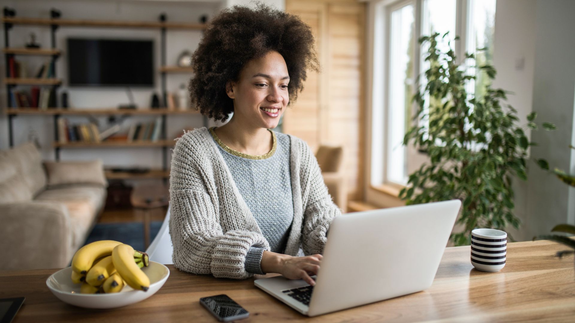 A person with curly hair sits at a wooden table typing on a laptop, with a bowl of bananas, a smartphone, and a striped mug beside them. There are bookshelves and plants in the background.